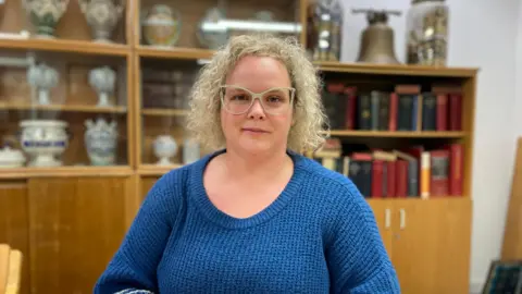 A portrait photo of Dr Caroline Copeland sitting at a table in a blue-knitted jumper. Behind her curly blond hair you can see shelves lined with jars from an old pharmacy with ornate lettering explaining the contents such as "opium" as well as research books