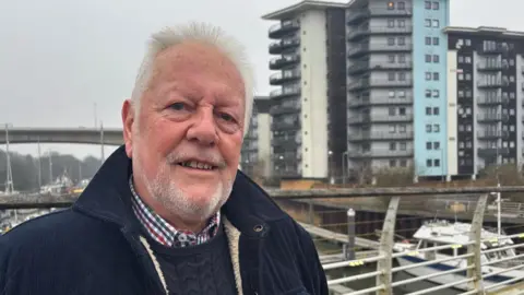 Man with white hair and white facial hair wearing a blue jumper, checked shirt, and navy jacket, smiling with his block of flats behind him