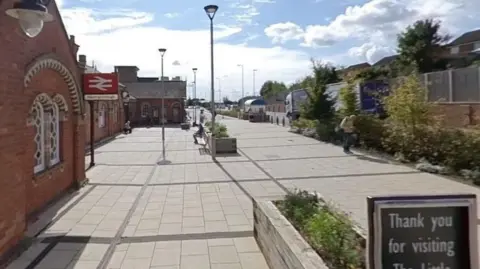 Station concourse made of grey paving slabs. A brick station building is visible to the left with a Wellingborough station sign.  A sign saying "Thank you for visiting..." is visible to the right, along with some small shrubs in a planter.