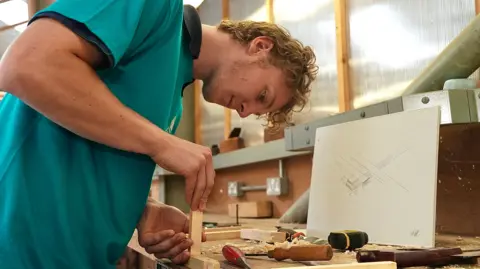 Martin Giles/BBC Phoenix has curly blonde hair, and he is working at his workbench. He is gluing one piece of wood to another. He is wearing a turquoise polo shirt with a navy blue polo shirt underneath.