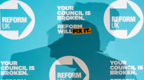 PA Media The shadow of Nigel Farage is cast onto a Reform UK campaign board after an election event in Doncaster in 2025. He is looking to the left, while another figure's shadow is looking right.