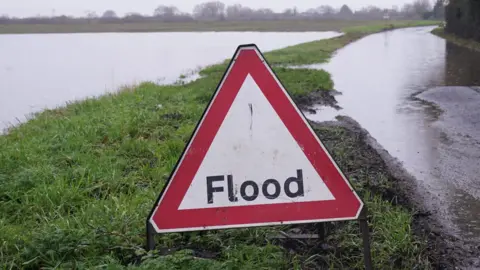 A road sign for flooding sits on an embankment next to a flooded road