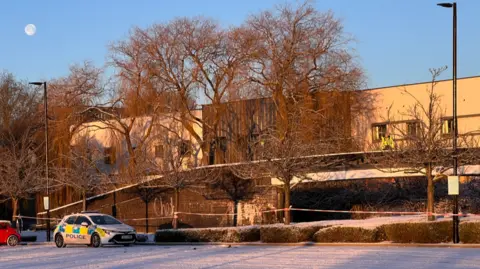 A police officer is standing on top of a bridge, with a police car parked to the side. There is red and white tape under the bridge
