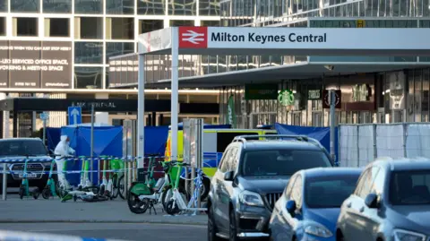 PA Media display outside the central Milton Keynes station. A police collar can be seen as a police car and a blue criminal tents. The forensic officer can be seen in a white suit walking inside the collar. The vehicles can be seen parked outside the station.