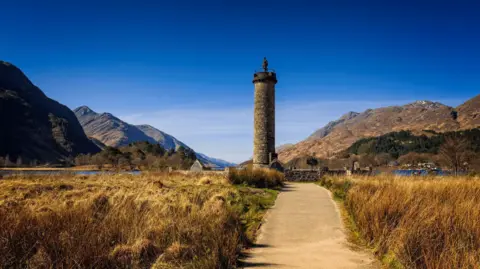 Glenfinnan Monument is a tower-like structure with a statue on top of it. The monument is surrounded by mountains and hills with Loch Shiel in the background. The scene is under a beautiful blue sky. 