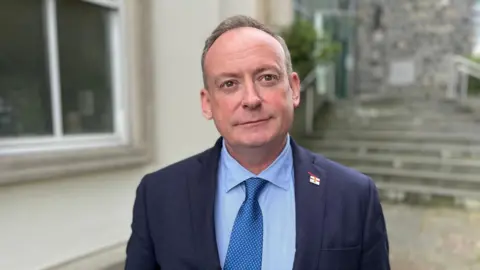 BBC A man wearing a blue shirt and a blue suit with a metallic blue tie looking towards the camera. On his left lapel he is wearing a Guernsey flag pin badge. 