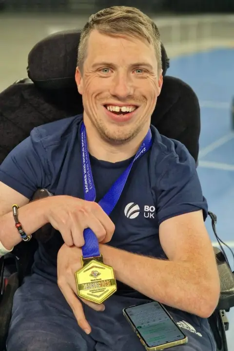 Claire Morrison David is sitting in a power chair with a big smile. He has short brown hair brushed to the side. David is holding a gold medal with a blue ribbon he has won.