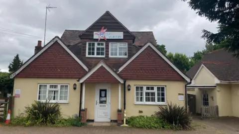 The front of a nursery building. The walls are yellow/beige, with brown brick on pointed parts of the roof. There is a sign in the centre of the roof reading "Capellas nursery"