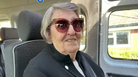Gavin Kermack An elderly woman with grey hair and sunglasses is sitting in a minibus. She is wearing a black jacket over a white top.