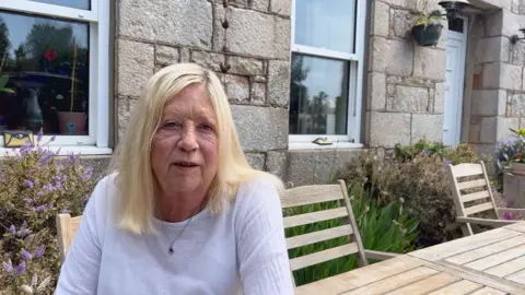 BBC Marina Mauger sat at a wooden table outside. She is wearing a white top and had short blonde hair. She is smiling at the camera. Behind her is a house, with a plants along the front of it.