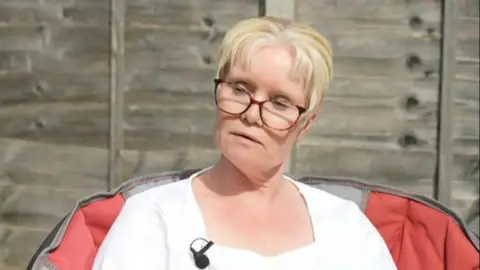 Lorraine pictured in white t-shirt. She has short blonde hair and large brown glasses. She is sat in front of a wooden fence. 