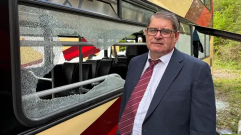 BBC Martin Spiller is standing next to a bus with smashed windows. He is wearing a dark grey suit with a claret-coloured tie with thin white stripes and a white shirt. He has grey hair and is wearing glasses. He looks upset.
