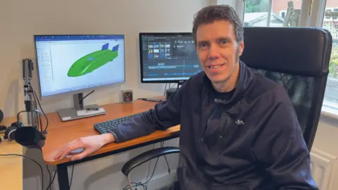 A man sits at desk on a chair, he is looking at the camera. Two monitors can be seen behind him. A prototype of a green remote-controlled car is seen on one screen and editing software on the other.