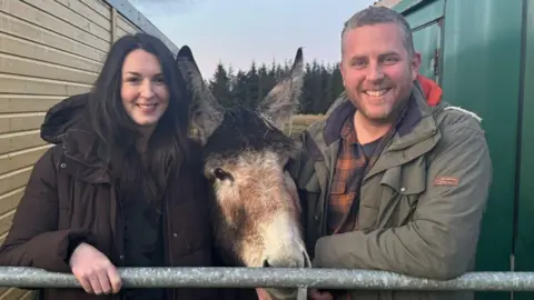 Macaulay College Una pareja sonriente parada junto a una puerta con un caballo entre ellos.