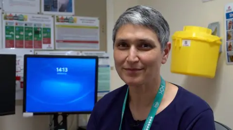 A woman in a lanyard with short grey hair smiles while sitting by a desk in a hospital.