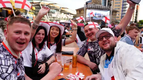 Owen Humphreys/PA A table of fans who are wearing headbands with little England flags sticking out from them or wearing England flag-themed cowboy hats.