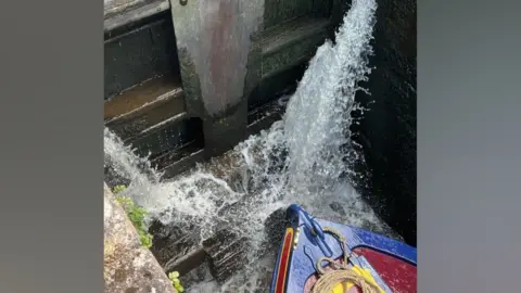 George Seward Water gushes in two streams through the gap between a pair of wooden lock gates. The bow of a red and blue narrowboat sits in the lock chamber below. Moss and small plants grow from the damp brickwork around the gates.
