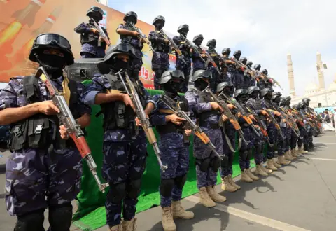 EPA Photograph showing a large formation of armed personnel standing in ranks on tiered platforms during a public display or parade. The individuals wear matching dark blue camouflage uniforms, black body armour, helmets with visors, face coverings, knee pads and tan military-style boots. Most hold rifles across their chests. The formation stretches diagonally across the frame, with personnel standing in multiple rows at different heights.