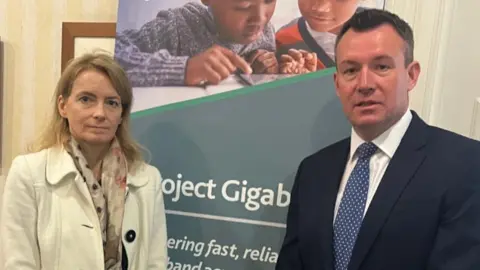 A woman with blonde hair and a cream jacket standing next to a man with short brown hair, a dark jacket and blue tie with a pale blue sign behind them