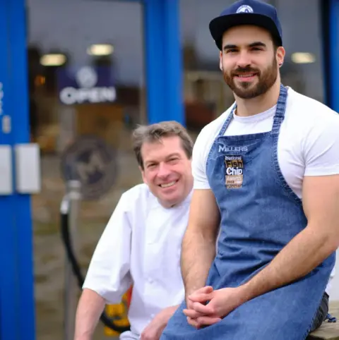 Millers Fish & Chips David and Nick wearing work uniforms sit outside a shop with bright blue doors. One person in front wears a denim apron with branding and a navy cap, along with a white T-shirt, seated casually with hands clasped. The person behind wears a white chef’s jacket. The shop’s glass door shows an ‘Open’ sign and reflections of the interior.”