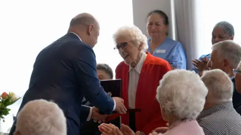 The Uzbekistan ambassador to the UK awarding Dulcie Le Breton The Order of Friendship at her care home. They are standing up 