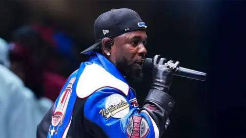 Getty Images Kendrick Lamar, performing at the Super Bowl in New Orleans, wearing a black leather glove, and singing into a black microphone, with a reversed black cap.