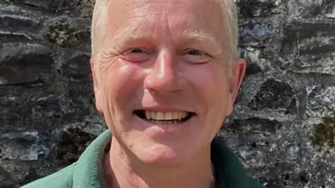 Quantock Hills Area of Outstanding Natural Beauty Andy smiles into the camera. He is standing in front of a grey stone wall. He has blonde hair and a green shirt. 