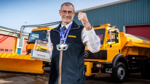 Econ Engineering Gary Lawson - a gritter driver - stands in front of two yellow Mercedes gritter vehicles. He is wearing a white shirt and navy tie and a blue fleece with the yellow branding 'ECON' on it. Gary has short grey hair and wears silver rimmed glasses. He is wearing two medals on lanyards and is holding an award which has a small yellow gritter on it. He is fist-pumping the air with joy.