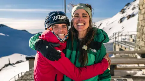 Jan Virt A close up of Fay Manners with her climbing partner Ella Wright. Both have their arms around each other and are smiling on top of snowy mountain set against a blue, slightly cloudy sky.