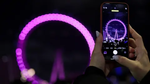 Reuters Tangan seorang wanita terlihat memegang ponsel pintar untuk mengambil gambar London Eye, yang dapat dilihat di layar ponsel dan di latar belakang gambar, sedikit tidak fokus.