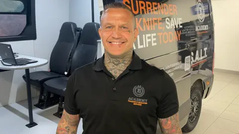 A man with short hair, a black shirt and tattoos on his arms and neck, smiles as he stands in front of a van with branding on it and a chairs, table and laptop inside.