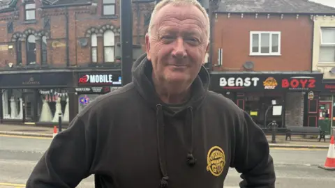 Andy Pritchard has a dark hoodie with a yellow badge.
