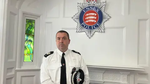 Henry Godfrey-Evans/BBC Stuart Hooper in a white shirt and black tie with a police hat under his arm and the Essex Police badge in the background