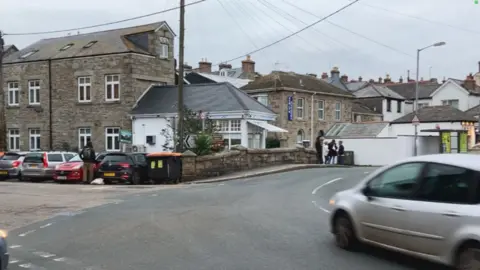 Newlyn. A silver car on a road bending to the left with car parks in bays on the left-hand side. There are stone buildings in the background