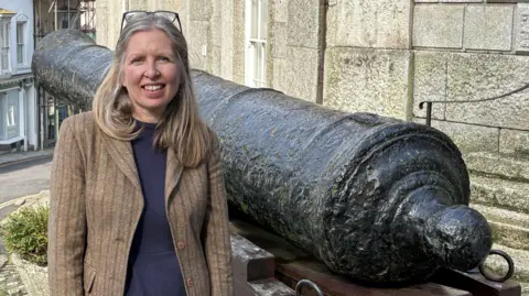 Annettee wears a dark blue top and light brown jacket and has light hair. She is standing in front of a statue of a replica cannon to her left - outside the Museum of Cornish Life entrance. 