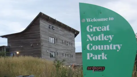 Lewis Adams/BBC A large building made of long wooden panels. It is three storeys high at the front, with a sloping, curved roof to the back, which has two storeys. In the foreground is a green sign that says "Welcome to Great Notley Country Park" in white font.