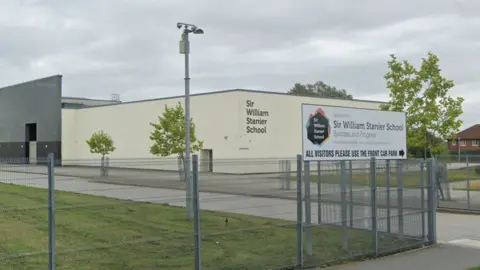 A large cream coloured square building reading "Sir William Stanier School" on the front. It is surrounded by grass and has small trees along a driveway outside. It has a white welcome sign at the front and metal fences.