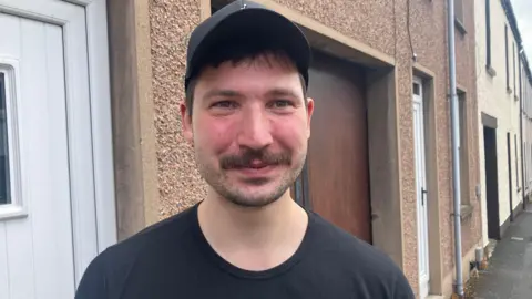 A man is smiling into the camera. He has brown hair and is wearing a black cap and a black top. He's standing on a street.