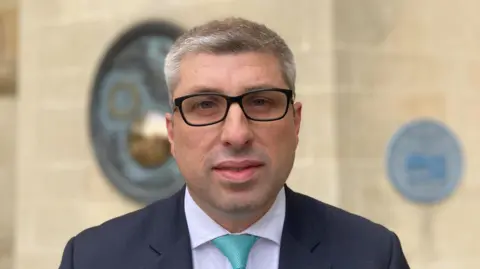 West Northants Council leader Mark Arnull wearing a navy suit, white shirt and turquoise tie