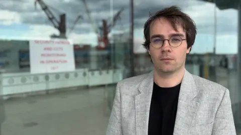 BBC/Jim Scott Councillor Paul Edgeworth, who is wearing circular black-framed glasses, a black shirt and a silver blazer, stands in front of the National Glass Centre. In the glass window behind him there is a sign which is warning people there is security operating, and is also reflecting a ship which is berthed opposite.