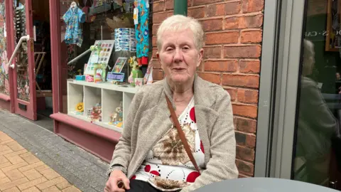 A woman is sitting at a small grey coffee table outside a row of shops. She looks straight into the camera. She has short white hair. She is wearing a white t-shirt patterned with red and brown geometric shapes. She is wearing a grey-brown cardigan over the t-shirt. She has a wooden walking stick in her right hand.