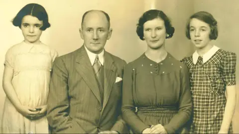 Dame Stephanie Shirley es una joven Dame Stephanie con una fotografía en blanco y negro con un vestido pálido en el lado izquierdo de sus padres y su hermana, y su familia también ha posado con expresiones neutrales en ropa inteligente.