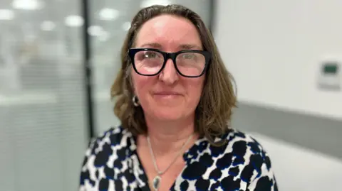Martin Giles/BBC Karen Thomas smiles at the camera. She has shoulder length brown hair and wears glasses and a black and white top. She has a silver necklace on with a green pendant in the middle of it. 