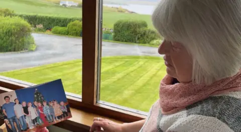 Kathleen next to a window overlooking her garden, holding a photograph of her 10 grandchildren. 