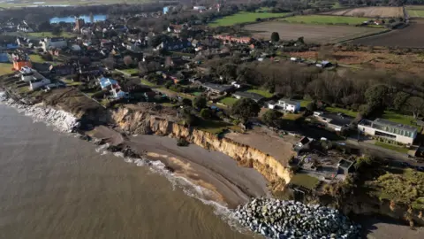 Joe Giddens/PA An aerial image looking inland from above the sea. A sandy cliff has been exposed by erosion, creating a bay. At either end, sea defences have been placed to try to protect the cliff. The rest of the village is visible behind, including a lake.
