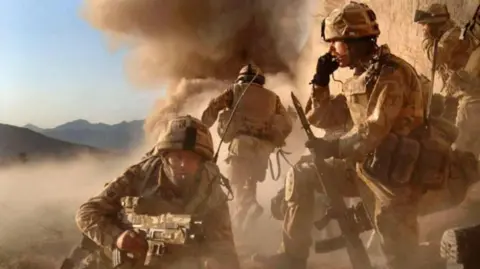 MOD (via PA) Soldiers in a battle scene in dusty Afghanistan. One soldier is crawling towards the camera, holding his rifle. Another is crouched on one knee, his rifle propped next to him, as an explosion in the background sends a plume of smoke into the sky.