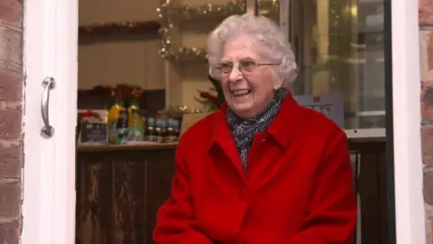 BBC An elderly woman wearing a red coat and black scarf stood in a doorway