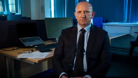 Yeti Television/BBC Cymru Wales Dorian Lloyd, former senior investigating officer at South Wales Police. Lloyd is a bald middle aged man who sits with his back to his desk. On his desk is papers, a pair of glasses and a computer. He wears a pinstripe shirt, black suit jacket and a black tie. 