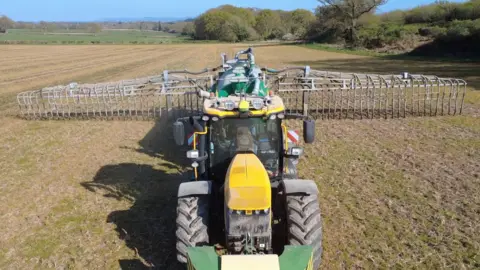 Frans de Boer An aerial view looking at a yellow tractor in the field. A green tank is on the back of the tractor. Long metal 'arms' come off the back of the tank and off the arms are multiple pipes going to the ground.