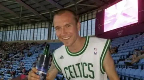 A man with short brown hair in a white vest with Celtics in green lettering on it, holding a bottle in a stadium with blue seating and a large number of people sat around him